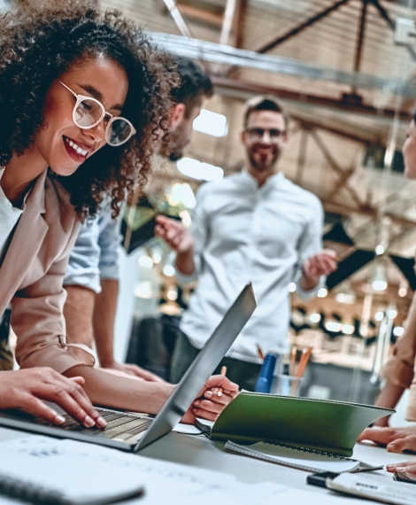 a woman works on a computer while her team is gathered around the table, offering insights and encouragement